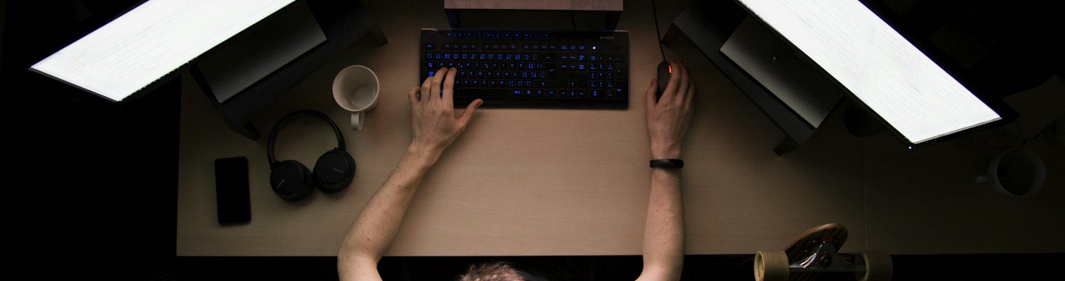 Overhead view of a person working at a desk with three bright monitors in a dark room.
