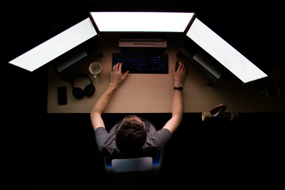Overhead view of a person working at a desk with three bright monitors in a dark room.